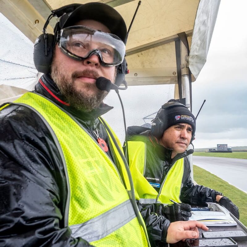 Two men in safety vests watch a race.